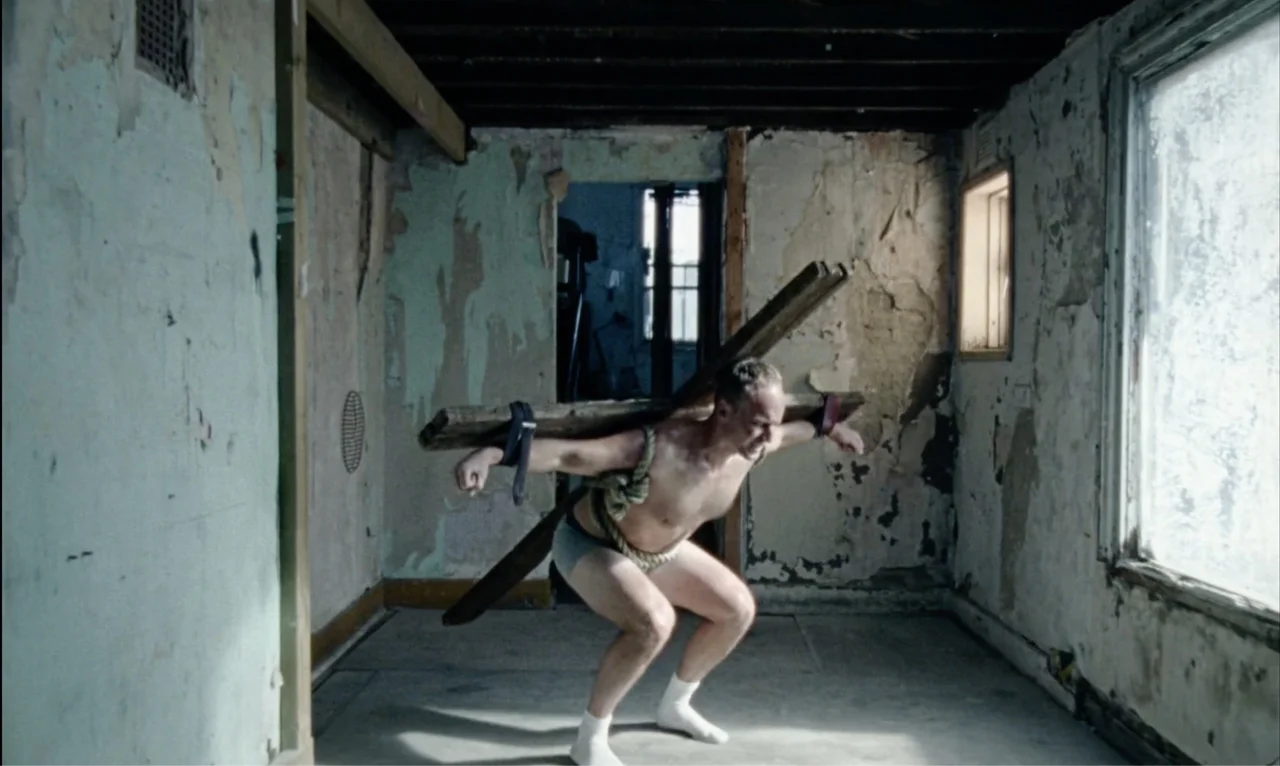 A figure carries a wooden crucifix through a derelict room.